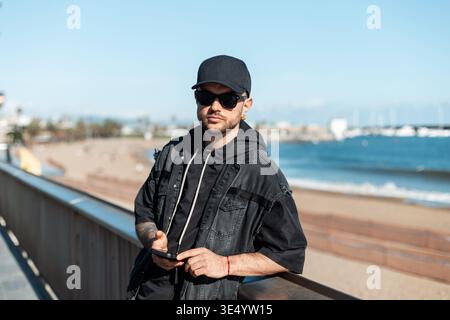 Stylish handsome fashion hipster man with sunglasses with cap in black denim jacket with hoodie holding smartphone and walking along beach in Barcelon Stock Photo