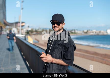 Happy handsome cool hipster man with black mockup cap with sunglasses in black denim clothes holding smartphone and standing on promenade in Barcelona Stock Photo