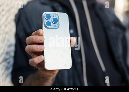 man holds a broken phone with cracks in his hand. The smartphone fell on the ground on the street and broke Stock Photo