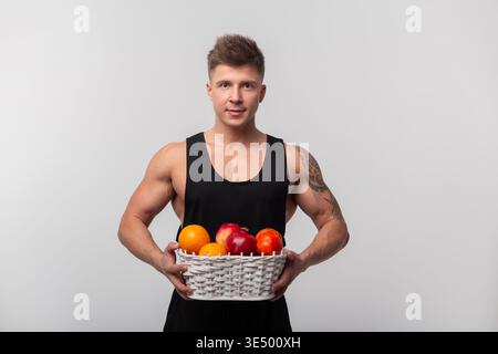 Handsome athletic man in black T-shirt holding basket of fruits on grey background. Diet, fruits and vitamins Stock Photo
