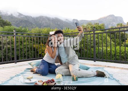 Couple with Asian woman taking selfie on rooftop patio sitting on blue picnic blanket with phone Stock Photo
