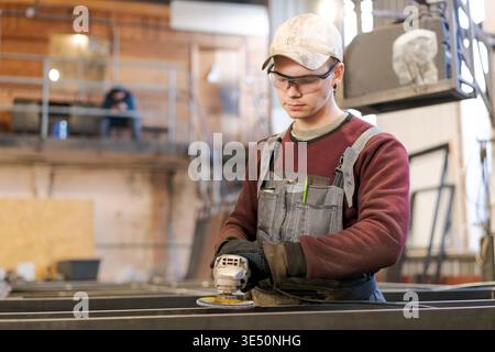 Concentrated young man in protective gear using angle grinder on steel construction in factory setting Stock Photo