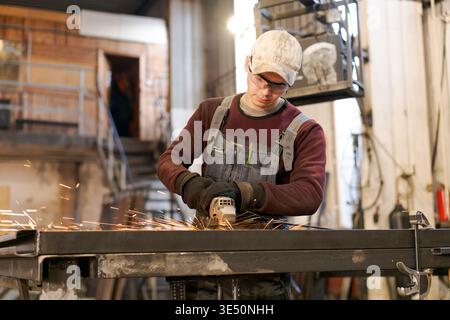 Young man in overalls and safety glasses using angle grinder on steel construction creating sparks in factory Stock Photo