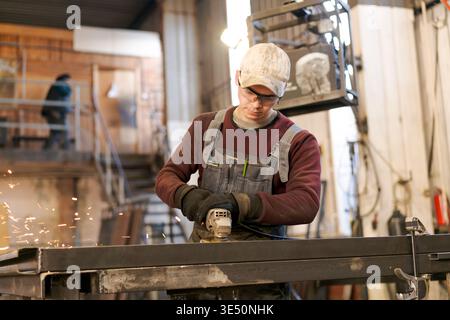 Young man in overalls and safety glasses using angle grinder creating sparks while working on steel frame Stock Photo