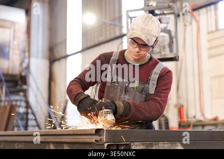 Focused young tradesman in protective eyewear using angle grinder creating sparks in manufacturing facility Stock Photo