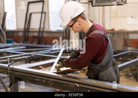 Young tradesman in white hardhat using angle grinder on metal construction with sparks in wide factory shot Stock Photo