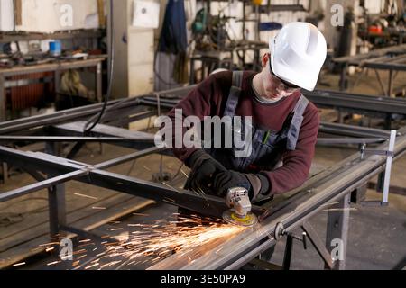 Young tradesman wearing white helmet using angle grinder on metal construction with sparks in wide workshop view Stock Photo