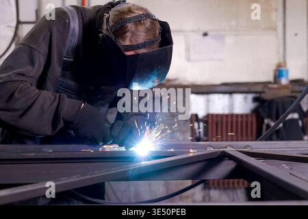 Experienced mature welder working on metal frame with bright welding arc and radiator visible in background of workshop Stock Photo