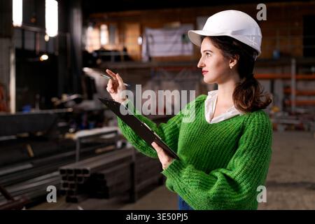Female engineer in safety helmet gestures with pen while supervising metal warehouse Stock Photo