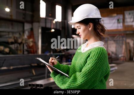 Woman in hard hat writes on clipboard while inspecting metal profiles at industrial warehouse Stock Photo