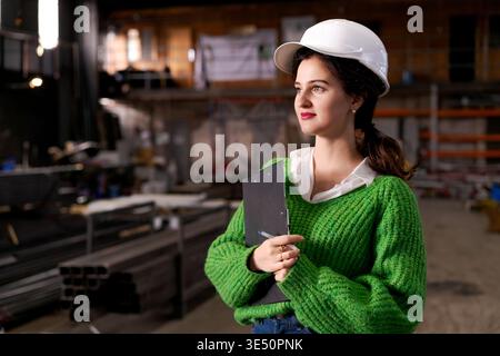Female engineer in safety helmet holds clipboard while surveying industrial storage facility Stock Photo