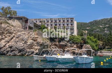 Small motorboats float on turquoise water near a rocky coastline below an abandoned seaside hotel. Lush green hills and blue sky, Corfu island, Greece Stock Photo