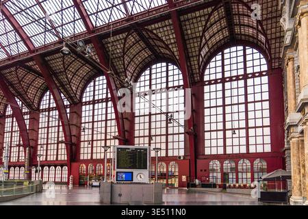 Monumental interior of Antwerp station featuring massive red iron arches and expansive glass windows. A train departure board stands in the foreground Stock Photo