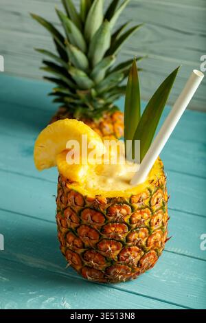 Fresh pineapple cocktail served in a hollowed pineapple with a straw and slice of pineapple on a turquoise wooden table Stock Photo