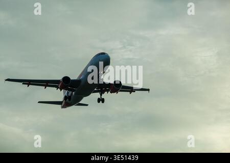 Commercial airplane flying through cloudy sky during sunset, showcasing landing gear extended and wings positioned for descent against a dramatic Stock Photo