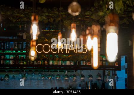 Neon sign reading "COCKTAILS" illuminated above a bar with hanging light bulbs and shelves stocked with various bottles and glassware in a modern Stock Photo