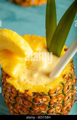 Fresh pineapple smoothie served in a hollowed pineapple shell with pineapple slices and green leaves, placed on a blue background with a straw Stock Photo