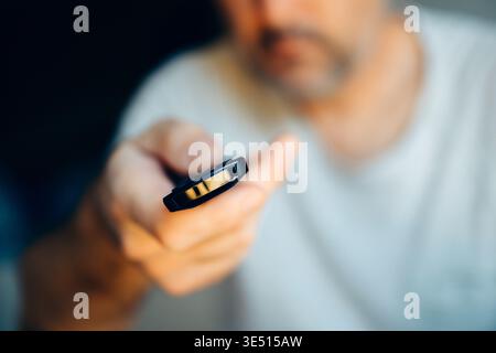 Close-up of man holding TV remote control with selective focus on buttons and fingers, face blurred in dark living room background Stock Photo