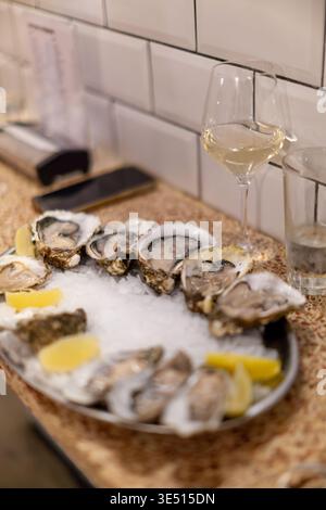 Fresh oysters served on ice with lemon wedges alongside glasses of white wine in a modern restaurant setting with tiled walls and wooden counter Stock Photo