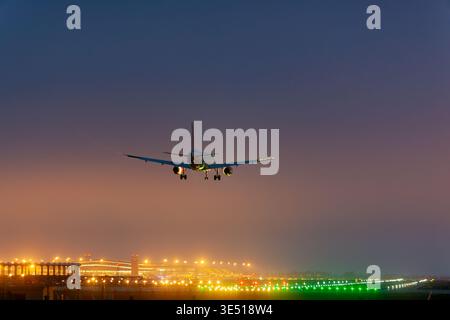 Airplane approaching landing at airport runway illuminated by colorful lights during twilight with foggy atmosphere and terminal buildings in the Stock Photo