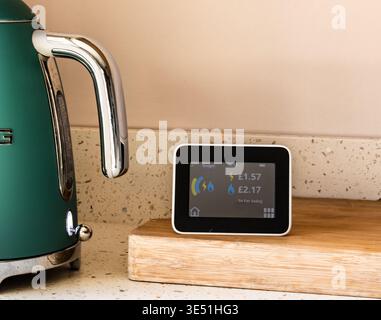 Smart electricity and gas meter monitor displaying energy usage and costs in a kitchen setting with a kettle nearby Stock Photo