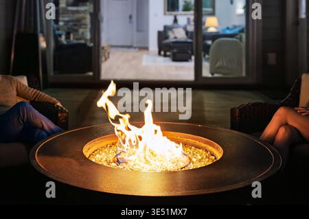 A young couple sit near a flaming firepit on an exterior garden deck with an illuminated room behind them at night. Stock Photo