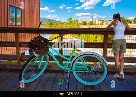 A young woman with a turquoise bicycle stops along the Cedar Street Bridge above Lake Pend Oreille Sand Creek in the small town of Sandpoint, Idaho. Stock Photo