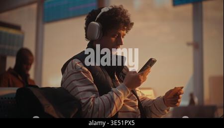 During Warm Golden Hour, Male Passenger Managing Travel Logistics. After Putting on Silver Headphones, Man Cross References Boarding Pass With Mobile App on Smartphone. Travel Apps, Airline Marketing. Stock Photo