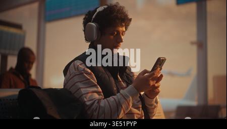 During Warm Golden Hour, Male Passenger Managing Travel Logistics. After Putting on Silver Headphones, Man Cross References Boarding Pass With Mobile App on Smartphone. Travel Apps, Airline Marketing. Stock Photo