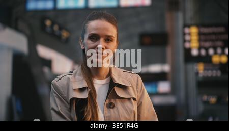 Young Woman in Beige Trench Stands Still and Smiles in Middle Busy Airport Departure Hall. While Other Passengers Walk Past in Motion Blur, Lady Remains Calm and Joyful, Looking Forward Her Journey. Stock Photo