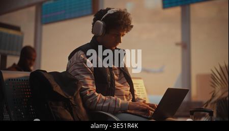 Young Man With Curly Hair Sits by Large Window in Airport Departure Lounge. Male Focused on Laptop Screen, Fingers Moving Rapidly Across Keyboard as Writes Code. Productivity of Modern Programmer. Stock Photo