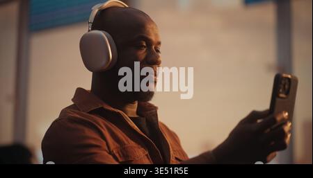 Black Man, Wearing Stylish Silver Over Ear Headphones and Overshirt, Engages in Video Chat on Smartphone. Sitting in Airport Departure Lounge, Man Smiles and Sharing Travel Excitement With Loved Ones. Stock Photo