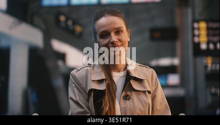Young Woman in Beige Trench Stands Still and Smiles in Middle Busy Airport Departure Hall. While Other Passengers Walk Past in Motion Blur, Lady Remains Calm and Joyful, Looking Forward Her Journey. Stock Photo