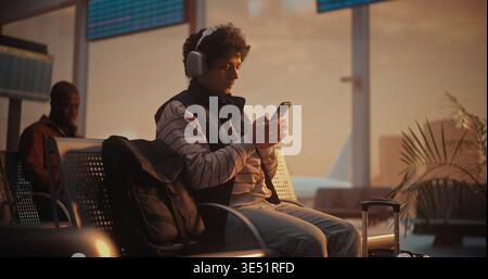 Focused Young Man in Striped Sweatshirt Putting on Large Over Ear Headphones. Male Uses Smartphone to Scan Yellow Paper Ticket, Instantly Pulling Up Digital Version on Screen to Verify Flight Details. Stock Photo