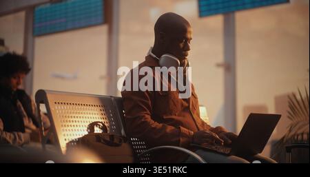 African American Man Sits on Terminal Bench, Deeply Focused on Laptop Screen While Waiting For Flight. Concept of Freelance Lifestyle, Corporate Travel, Mobile Office Solutions, Global Connectivity. Stock Photo
