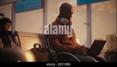 African American Man Sits on Terminal Bench, Deeply Focused on Laptop Screen While Waiting For Flight. Concept of Freelance Lifestyle, Corporate Travel, Mobile Office Solutions, Global Connectivity. Stock Photo