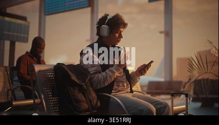 Young Man With Curly Hair Enjoys Healthy Snack While Waiting For Flight. Male Sits in Airport Terminal, Wearing Large Silver Over Ear Headphones, Taking Bite of Banana While Focused on Smartphone. Stock Photo