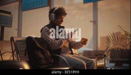 Focused Young Man in Striped Sweatshirt Putting on Large Over Ear Headphones. Male Uses Smartphone to Scan Yellow Paper Ticket, Instantly Pulling Up Digital Version on Screen to Verify Flight Details. Stock Photo