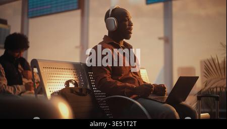 Sitting in Departure Lounge, Black Professional Engages in Business Video Call on Laptop. Man Wears Headphones For Clear Audio, Speaking Confidently While Warm Golden Hour Light Fills Terminal. Stock Photo