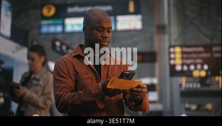 African American Man Stands In Brightly Lit Airport Terminal, Using Smartphone to Scan Digital Boarding Pass. Male Carefully Reviews Flight Details, Gate Numbers, and Departure Time on His Screen. Stock Photo