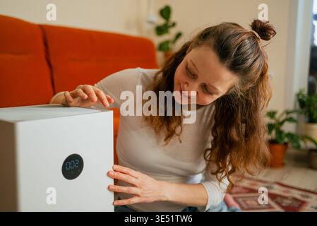 Woman monitoring and interacting with a modern white air purifier at home, promoting clean breathing and well-being in a comfortable living space with advanced smart technology Stock Photo