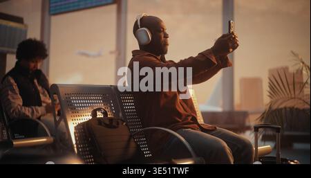 African American Man Sits in Airport Waiting Area and Captures Moment. Male Holds Smartphone Up With One Hand to Take Selfie. Staying Connected With Friends or Social Media During Flight Layover. Stock Photo