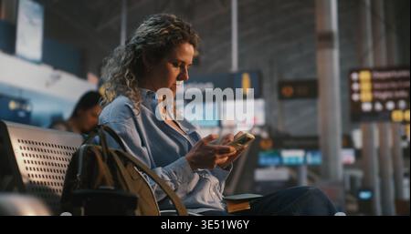 Woman Sits in International Airport Terminal, Using Smartphone Scan QR Code on Boarding Pass. Female Verifies Flight Data While Enjoying Cup of Coffee. Concept of Paperless Travel, Digital Check In. Stock Photo