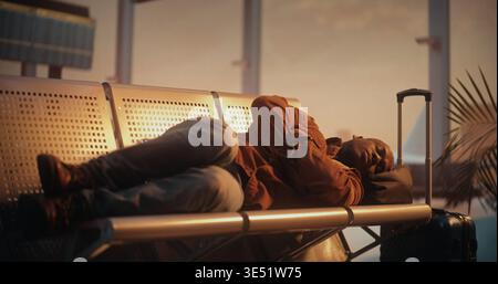 Weary African American Male Passenger in Overshirt Attempts to Get Some Rest While Waiting For Boarding Call. Man Curled Up on Airport Benches. Concept of Struggle to Find Comfort in Public Spaces. Stock Photo
