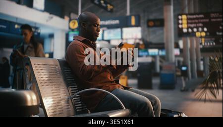 Black Male, Pulling Suitcase Enters Airport Waiting Area. Man Finds Seat and Uses Smartphone Scan Boarding Pass, Glances Between Device and Overhead Flight Information Boards Confirm at Correct Gate. Stock Photo