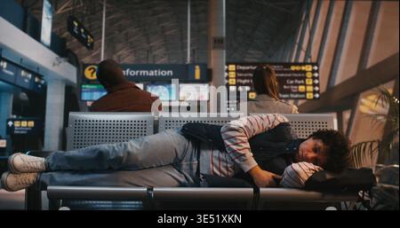 Exhausted Traveler Adjusts Position on Cold Metal Airport Chairs, Tucking Black Backpack Under Head For Comfort. After Long Wait in Terminal, Man Closes Eyes to Catch Some Sleep During Flight Delay. Stock Photo