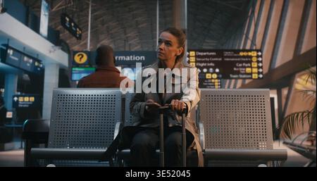 Stylish Female Traveler Sits in Departure Lounge With Suitcase. Woman Checks Smartphone, Looks at Wristwatch to Verify Time, and Finally Glances Around Terminal With Calm Smile. Travel Punctuality. Stock Photo
