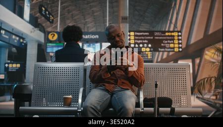 Young Black Man Arrives at Airport Waiting Area, Sets Down Luggage, Immediately Checks Smartphone. Modern Habit of Staying Connected, Checking Flight Updates, Chatting With Friends Before Departure. Stock Photo