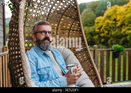 Man is enjoying cup of warm tea, coffee, having relaxing moment at home, sitting in hanging chair on patio. Stock Photo