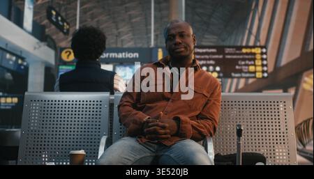 Black Man Sits in Airport Lounge, Using Smartphone Scan QR Code on Ticket. While Waiting Flight, Male Traveler Drinks Cup of Coffee, Calmly Double Checks Departure Details. Modern Travel Technology. Stock Photo